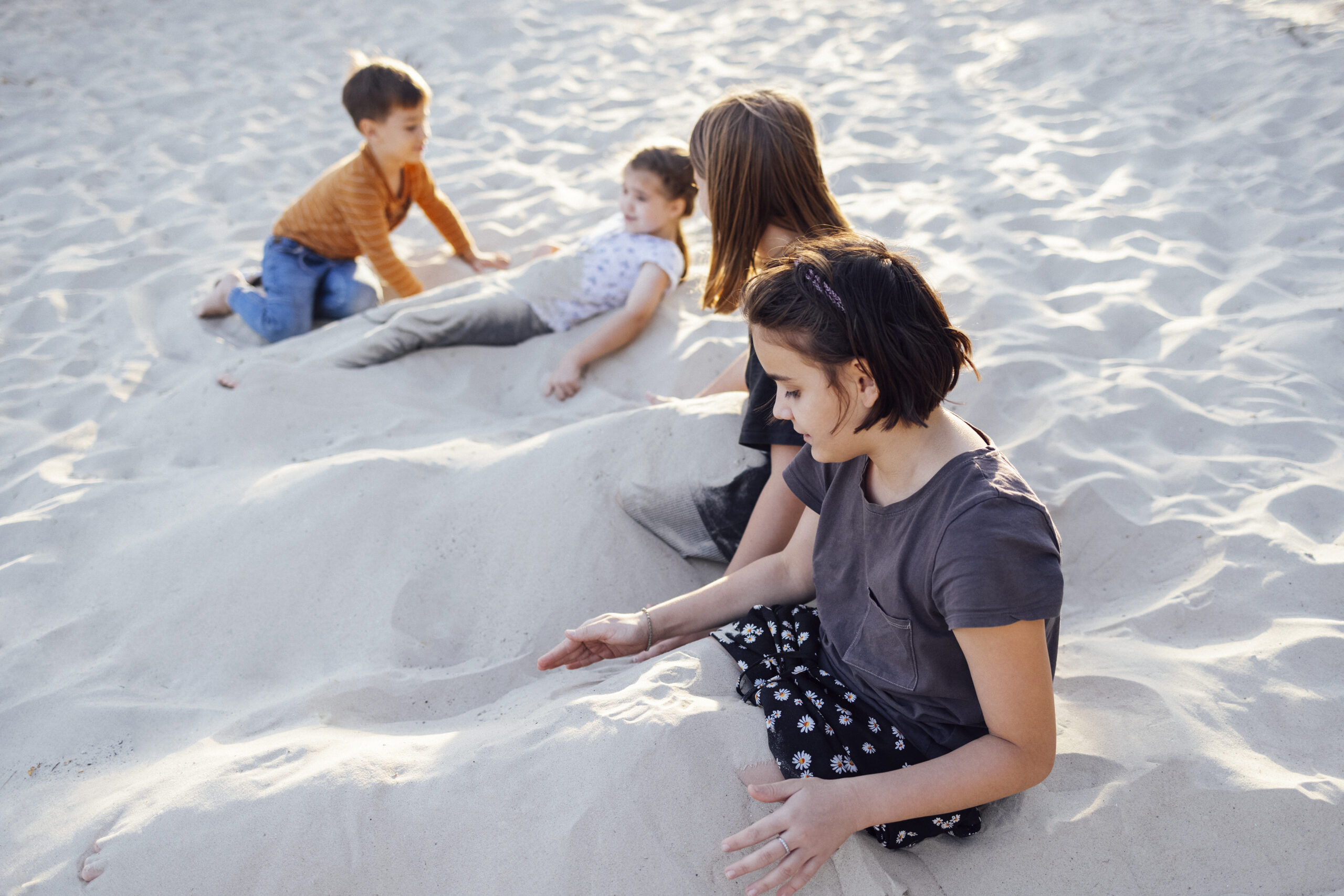 Four joyful children in casual clothes play on beach. Funny girls pour sand on themselves. Smiling sisters dig their legs into sand. Cute kids have fun near sea. Family vacation and happy childhood.